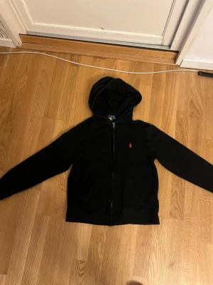Svart hoodie med dragkedja och huva - Säljer en svart hoodie med dragkedja och huva. Den har två stora fickor framtill och en liten röd logga broderad på bröstet. Hoodien är i mjukt material och har ribbade muddar vid ärmar och nederkant. Perfekt för en chill och avslappnad stil.