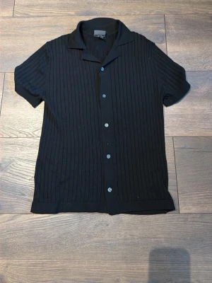 Svart ribbad piké med knappar - Snygg svart piké med ribbad struktur. Pikén har klassisk krage och knäppning framtill. Perfekt för en stilren look och enkel att matcha med olika outfits. Storlek: XS, Passfrom: Regular fit