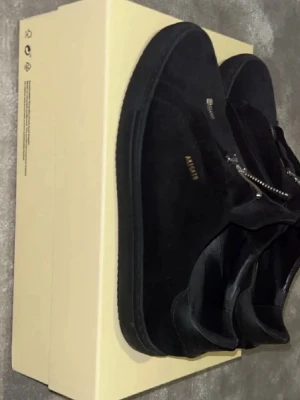 Arigatos med dragkedja - Stilrena svarta sneakers från Axel Arigato i mocka med silverfärgad dragkedja framtill. Låg modell med diskret logga i guld på sidan. Perfekta för dig som gillar minimalistisk och modern design. Storlek 44 