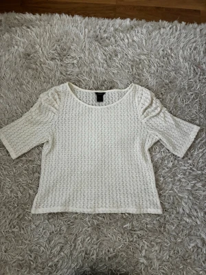 Vit stickad topp med puffärm - Söt vit stickad topp med strukturmönster och puffade korta ärmar. Toppen har rund halsringning och är croppad i modellen, perfekt för en trendig look. Materialet känns mjukt och luftigt, vilket gör den skön att bära under varmare dagar men även nu i höst.