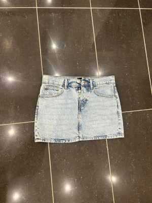 Ljusblå jeanskjol med knappar, tagit på mig 1 gång - Säljer en ljusblå jeanskjol i klassisk modell med knappgylf och fem fickor. Kjolen har bälteshällor och är i en kort längd, perfekt till sommaren. Materialet är denim och kjolen har en avslappnad passform.