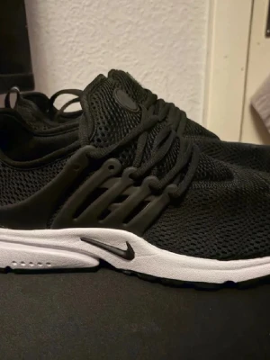 Svarta Nike sneakers med vit sula - Svarta sneakers från Nike med mesh-ovandel och vit sula. Skorna har en sportig design med svarta snören och plastdetaljer på sidorna för extra stöd. Perfekta för en stilren och bekväm look.