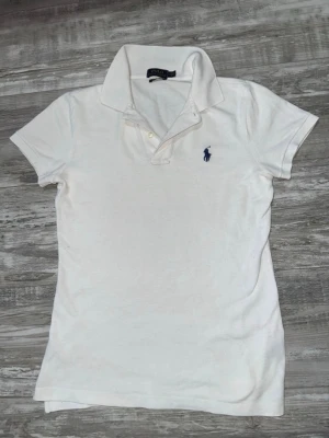 Vit pikétröja från Polo Ralph Lauren - Klassisk vit pikétröja från Polo Ralph Lauren med korta ärmar och blå broderad logga på bröstet. Tröjan har krage och knäppning med två knappar. Perfekt till jeans eller shorts för en clean och tidlös look.