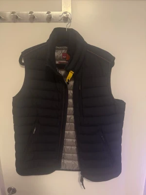 Svart dunväst från Parajumpers - Snygg svart dunväst från Parajumpers med quiltad design och hög krage. Västen har dragkedja framtill, två sidofickor med dragkedja och en gul detalj vid halsen. Insidan är ljusgrå och materialet är lätt och värmande. Den är helt ny använd max 5 gånger.