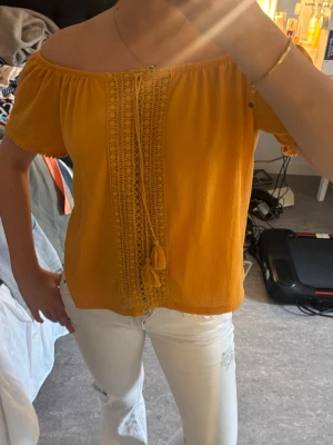 Gul offshoulder topp med spetsdetaljer - Snygg gul offshoulder topp med korta ärmar och dekorativa spetsdetaljer framtill. Toppen har snörning med tofsar och är gjord i ett lätt, luftigt material som passar perfekt till sommaren. Superskön.