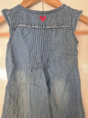 Blå jeansklänning med hjärtdetaljer - Söt blå jeansklänning i mjukt denimtyg med röda hjärtan broderade på fickorna och ryggen. Klänningen har korta ärmar, knappar framtill och volangkant i rosa tyg längst ner. Smockdetaljer framtill ger en fin passform och extra stil.i storlek 92 