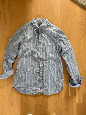 Blåvit randig skjorta Polo Jeans Co. - Snygg blå skjorta med vita vertikala ränder från Polo Jeans Company. Skjortan har klassisk krage med knappar, bröstficka och långa ärmar. Tillverkad i bomull för en bekväm känsla och stilren look. Ralph Lauren