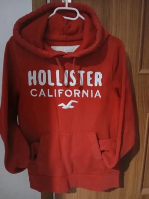Röd hoodie från Hollister - Röd hoodie från Hollister med vit text 'Hollister California' och logga framtill. Klassisk känguruficka, dragsko i huvan och mjukt material. Perfekt för en avslappnad och cool stil.