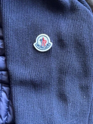 Mörkblå dunväst från Moncler - Snygg mörkblå dunväst från Moncler med quiltad framsida och stickade sidor. Västen har dragkedja framtill och klassisk Moncler-logga på bröstet. Perfekt för lager-på-lager och ger en sportig vibe till din outfit.