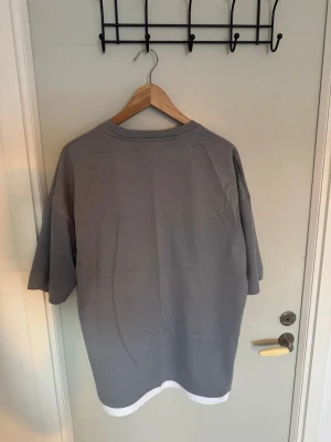 Grå oversized t-shirt med vit kant - Säljer en grå oversized t-shirt med vit kant längst ner. T-shirten har rund halsringning och korta ärmar. Perfekt för en avslappnad och trendig stil. Materialet känns mjukt och skönt, troligtvis bomull.