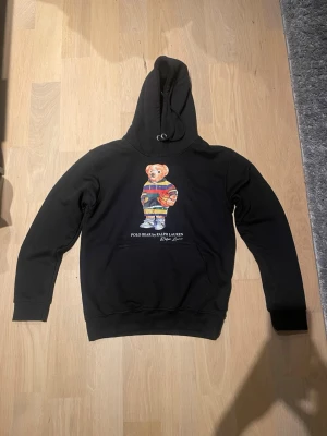 Svart hoodie Polo Bear Ralph Lauren - Säljer en svart hoodie från Ralph Lauren med den ikoniska Polo Bear-trycket på bröstet. Hoodien har huva med dragsko och ribbade muddar vid ärmar och nederkant. Materialet ser ut att vara mjukt och bekvämt, perfekt för en avslappnad stil.