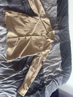 Beige overshirt från C.P. Company - Snygg beige overshirt i manchestertyg från C.P. Company. Klassisk krage, knappstängning framtill och en bröstficka. Unik detalj med C.P. Company-logga på ärmen. Perfekt lager på lager-plagg för en trendig look.
