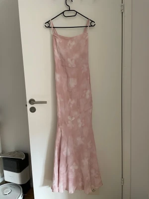 ADOORE venlo dress stl 34 - Säljer en elegant långklänning i ljusrosa med vita blommor. Klänningen har smala axelband, figurnära passform upptill och faller snyggt nedtill. Materialet ser ut att vara mjukt och glansigt, troligen satin eller polyester. Perfekt för sommarens tillfällen!