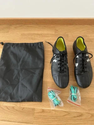 Under Armour Speedform fotbollsskor - Snygga svarta Under Armour Speedform fotbollsskor med vita detaljer och logga på sidan. Skorna har snörning, gult innerfoder och kommer med extra gröna skosnören samt en svart skopåse. Perfekta för dig som vill ha stilrena och lätta skor på planen.