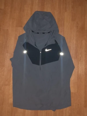 Grå Nike Miller - Snygg grå vindjacka från Nike med svart panel framtill och reflekterande detaljer. Jackan har huva, halv dragkedja och en sportig look. Perfekt för blåsiga dagar och outdoor-aktiviteter. Materialet känns lätt och smidigt.