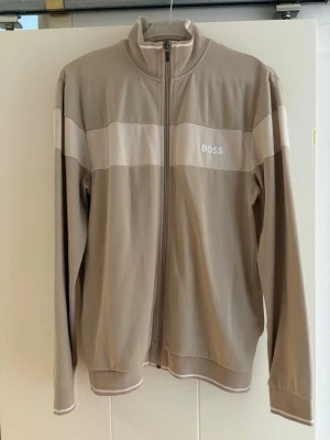 Beige ziptröja från BOSS - Snygg beige ziptröja från BOSS med vit logga på bröstet och breda vita ränder över bröst och armar. Tröjan har hög krage, hel dragkedja och ribbade muddar. Perfekt för en avslappnad och stilren look.