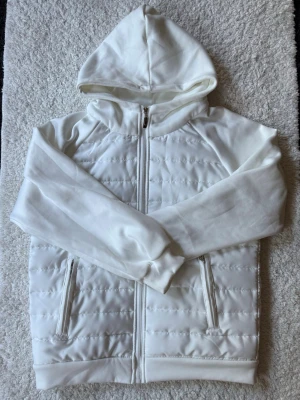 Hybrid jacka  - Snygg vit vindjacka med quiltad framsida och dragkedja hela vägen. Jackan har två fickor med dragkedja och en mjuk huva. Materialet är en mix av slät polyester och vadderad front för extra värme. Perfekt för dig som gillar stilren och enkel design. Mått: armhåla till handled 46cm. Midjan: 52cm. Axel till midjan 60cm.