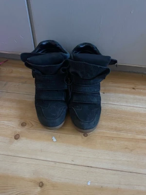 Svarta Wedges Sneakers - Svarta sneakers med högt skaft och flera breda kardborreband. Skorna har en chunky design med vadderad plös och krage samt en brun sula. Materialet ser ut att vara mocka eller liknande tyg. Perfekta för en streetwear-look.