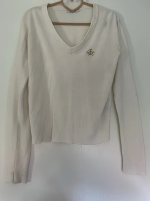 Vit stickad tröja med v-ringning - Klassisk vit stickad tröja med djup v-ringning och ribbade muddar. Tröjan har ett diskret broderat emblem på bröstet och är långärmad. Perfekt att matcha med jeans eller kjol för en clean look.