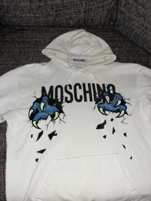 Vit Moschino hoodie med klor-print - Säljer en vit hoodie från Moschino med stort svart logotryck och coolt motiv av blå monsterklor som river genom tyget. Hoodien har huva med dragsko och magficka. Perfekt för dig som vill sticka ut med en unik och trendig design.