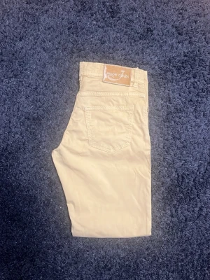 Beige chinos från Jacob Cohen - Snygga beige chinos från Jacob Cohen i mjukt bomullsmaterial. Klassisk femficksmodell med normal midja och raka ben. Perfekta för en clean och stilren look. Diskret logga på bakfickan och patch i läder baktill. Har gått bort lite av kosa skinnet.