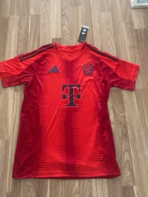 Röd Bayern München matchtröja - Säljer en röd Bayern München matchtröja från Adidas med svarta detaljer och klubbmärke på bröstet. Tröjan har korta ärmar, V-ringning och tre svarta ränder på axlarna. Perfekt för fotbollsträning eller att bära till vardags.