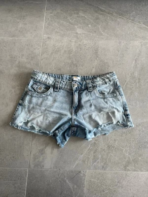 Ljusblå jeansshorts med broderi - Säljer ett par ljusblå jeansshorts med råa kanter och snygga broderade detaljer på bakfickorna. Shortsen har fem fickor, bälteshällor och knappgylf. Perfekta för sommaren och ger en avslappnad vibe.