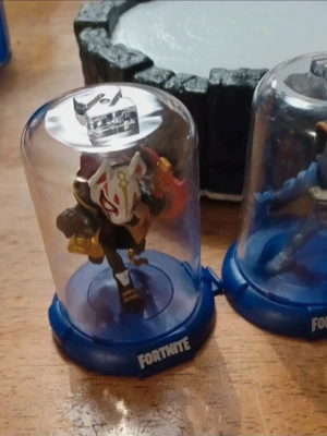 Fortnite spel med platta -  Fortnite  i genomskinliga plastkapslar med blå botten. Ena figuren har en vit rödmask och gulddetaljer, den andra är blå med rustning. Perfekt för Fortnite-fans 