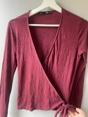 Vinröd omlottopp med knyt från 157 - Säljer en vinröd omlottopp från 157 i storlek S. Toppen har långa ärmar, v-ringning och knyt i sidan. Perfekt att styla med jeans eller kjol för en trendig look.