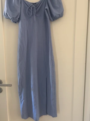 Blå långklänning från Gina Tricot - Säljer en ljusblå långklänning från Gina Tricot med puffärmar och snörning framtill. Klänningen har en mjuk och skön bomullskänsla och är perfekt för sommarens alla tillfällen. Stilren och enkel design med en feminin touch.