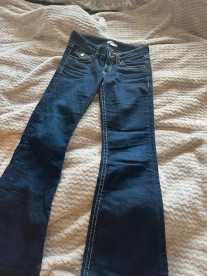 Mörkblå jeans - Säljer lillasyrrans gamla mörkblå bootcut par jeans med vita kontrastsömmar och klassisk femficksdesign. Jeansen har låg midja och är tillverkade i stretchigt denimtyg för skön passform. Perfekta för dig som gillar retrovibbar och vill ha ett par jeans som sticker ut. Storlek 158 och inga defekter 