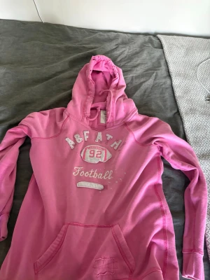 Rosa hoodie från Abercrombie & Fitch - Snygg rosa hoodie från Abercrombie & Fitch med tryck framtill och texten 'A&F ATH 92 Football'. Hoodien har huva med dragsko och en stor magficka. Perfekt för en avslappnad och sportig stil. Tillverkad i mjukt bomullsmaterial.