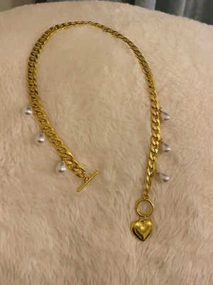 Guldigt halsband med pärlor och hjärta - Snyggt halsband i guldtonad rostfritt med vita pärlor längs kedjan och ett hängande hjärta som detalj. Kedjan är chunky och har ett T-lås framtill. Perfekt accessoar för att lyfta din outfit med en trendig touch.