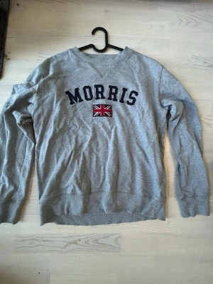 Grå sweatshirt från Morris - Snygg grå sweatshirt från Morris med broderad text och brittisk flagga på bröstet. Klassisk rund halsringning och ribbade muddar vid ärmslut och nederkant. Perfekt för en avslappnad och stilren look.
