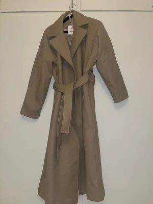 Beige lång kappa med knytskärp - Helt ny, stilren beige kappa i lång modell med bred krage och knytskärp i midjan. Kappan har raka ärmar och en klassisk siluett som ger en elegant look. Perfekt för dig som gillar minimalistisk och tidlös stil, storlek S/M
