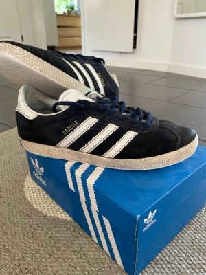 Adidas Gazelle marinblå sneakers - Klassiska Adidas Gazelle sneakers i marinblå mocka med vita detaljer och de ikoniska tre ränderna på sidan. Låg modell med vit sula och mörkblå snören. Adidas-logga på plösen och guldfärgad Gazelle-text på sidan. Perfekta för en stilren och laidback look. Storlek = 38 2/3 Hör gärna av er vid frågor! 
