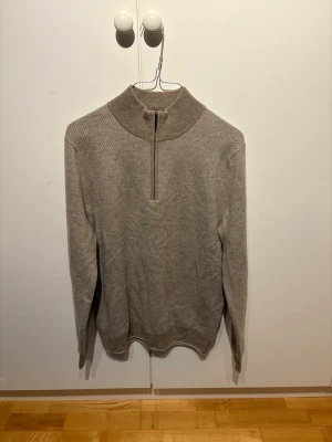 Beige stickad tröja - Säljer en stilren beige stickad tröja med hög krage och halv dragkedja framtill. Tröjan har långa ärmar och ribbade muddar vid ärmslut och nederkant.