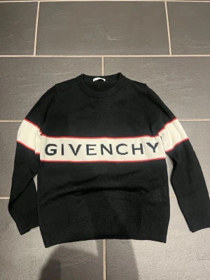 Givenchy svart stickad tröja - Svart stickad tröja från Givenchy med vit panel och röd linje över bröstet. Stor Givenchy-logga framtill, rund hals och ribbade muddar. Tröjan har lång ärm och är perfekt för dig som gillar exklusiv och trendig streetwear