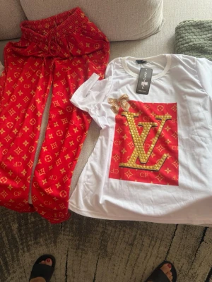 Röd och vit pyjamas med guldtryck - Säljer ett set med pyjamas i två delar: röda byxor med guldfärgade mönster och en vit t-shirt med stort guldigt LV-tryck och pärldetaljer. T-shirten har rund hals och kort ärm, byxorna har resår i midjan. Materialet ser ut att vara mjukt och bekvämt, perfekt för hemmamys.