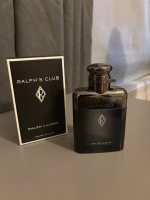 Ralph's Club Eau de Toilette - Ralph's Club Eau de Toilette från Ralph Lauren i en elegant, mörkgrå glasflaska med metallisk finish och stilren logotyp på framsidan. Flaskan har en modern, maskulin design och kommer med originalkartong i svart med vit text.