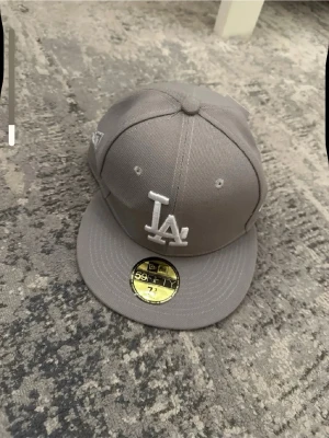 Grå LA-keps från New Era 59FIFTY - Snygg grå keps från New Era 59FIFTY med LA-logga framtill i vitt. Klassisk flat brim och broderade detaljer. Perfekt för dig som gillar streetstyle och sportiga vibes. Kepsen är i ett stadigt material och har märkets ikoniska sticker kvar på skärmen.