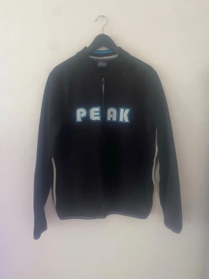 Svart ziptröja från  PEAK performance  - Svart ziptröja med blått och vitt PEAK-tryck på bröstet. Långärmad modell med dragkedja framtill och blå detaljer längs sidorna. Perfekt för en sportig och avslappnad stil.