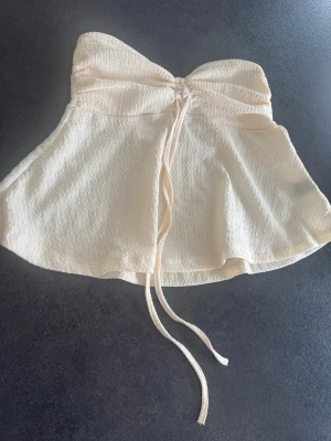 Beige bandeau tubtopp med snörning - Supersöt beige bandeau tubtopp med snörning framtill. Toppen är ärmlös och har en lätt utsvängd form nedtill. Perfekt för varma dagar och enkel att styla med jeans eller kjol.