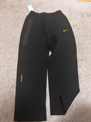 Svarta Nike mjukisbyxor med gul logga - Säljer ett par svarta mjukisbyxor från Nike med gul logga och textdetaljer. Byxorna har bred passform, dragsko i midjan och en dragkedjeförsedd ficka på sidan. Perfekta för chill dagar eller streetwear-stil.