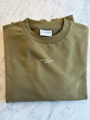 Olivgrön sweatshirt från Drôle de Monsieur - Säljer en olivgrön sweatshirt från Drôle de Monsieur med texten 'NOT FROM PARIS MADAME' på bröstet. Tröjan har rund hals, ribbade muddar vid ärmslut och nederkant samt är gjord i mjuk bomull. Perfekt för en avslappnad och stilren look. Tröjan är oanvänd. Nypris 1400 SEK.