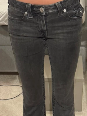 Mörkgrå bootcut jeans med detaljer - Säljer ett par mörkgrå bootcut jeans med snygga sömmar och silverfärgade knappar. Jeansen har fem fickor och en lätt utsvängd passform nedtill. Perfekta för dig som gillar en klassisk men trendig look.