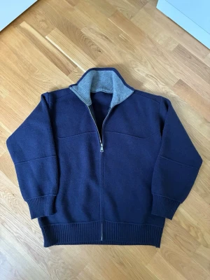 Stickad ziptröja - Mörkblå stickad tröja med dragkedja framtill och hög krage med grå insida. Tröjan har ribbade muddar vid ärmslut och nederkant, samt en avslappnad passform. Perfekt för lager på lager och håller dig varm under kyliga dagar.