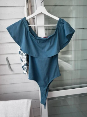 Blå off shoulder baddräkt Guess XS - Trendig blå baddräkt från Guess i off shoulder-modell med bred volang runt axlarna. Tillverkad i ett mjukt, stretchigt material som sitter snyggt på kroppen. Perfekt för strandhäng eller poolparty.