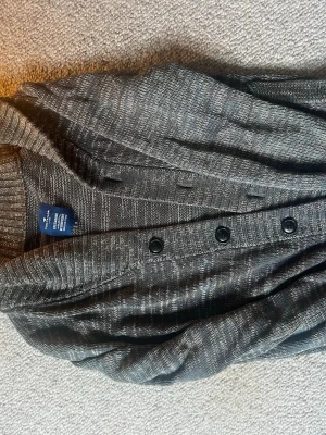 Gråmelerad kofta från Tom Tailor - Snygg gråmelerad kofta från Tom Tailor med knappar framtill och ribbade muddar. Koftan har en avslappnad passform och är stickad i mjukt material, perfekt för lager på lager. Klassisk design som funkar till det mesta.