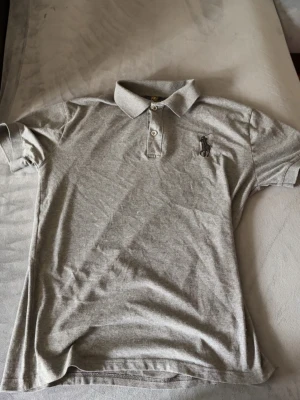 Grå pikétröja från Polo Ralph Lauren - Klassisk grå pikétröja från Polo Ralph Lauren med korta ärmar, krage och knäppning med tre knappar. Tröjan har den ikoniska broderade loggan på bröstet och är tillverkad i mjukt bomullsmaterial.